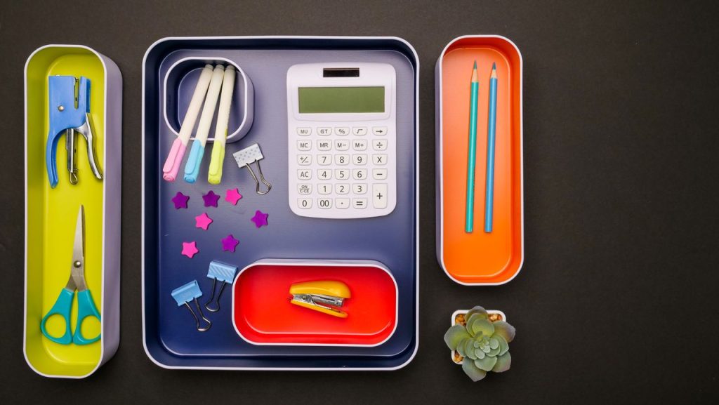 Flat lay of organized desk accessories in colorful trays including calculator, pens, and succulent plant.